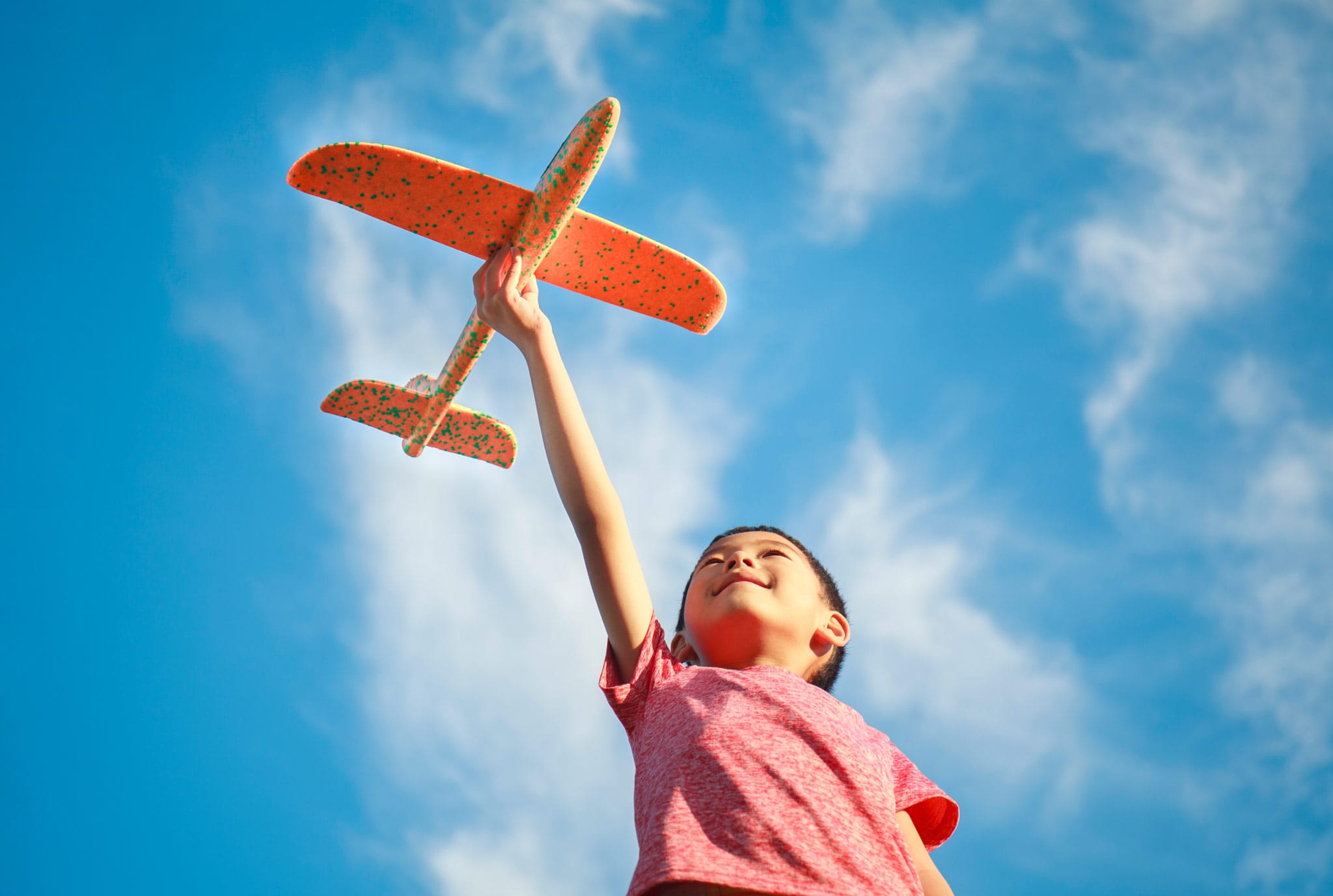 A child in a red shirt holds an orange toy plane aloft against a vibrant blue sky with wispy clouds, conveying joy and imagination.