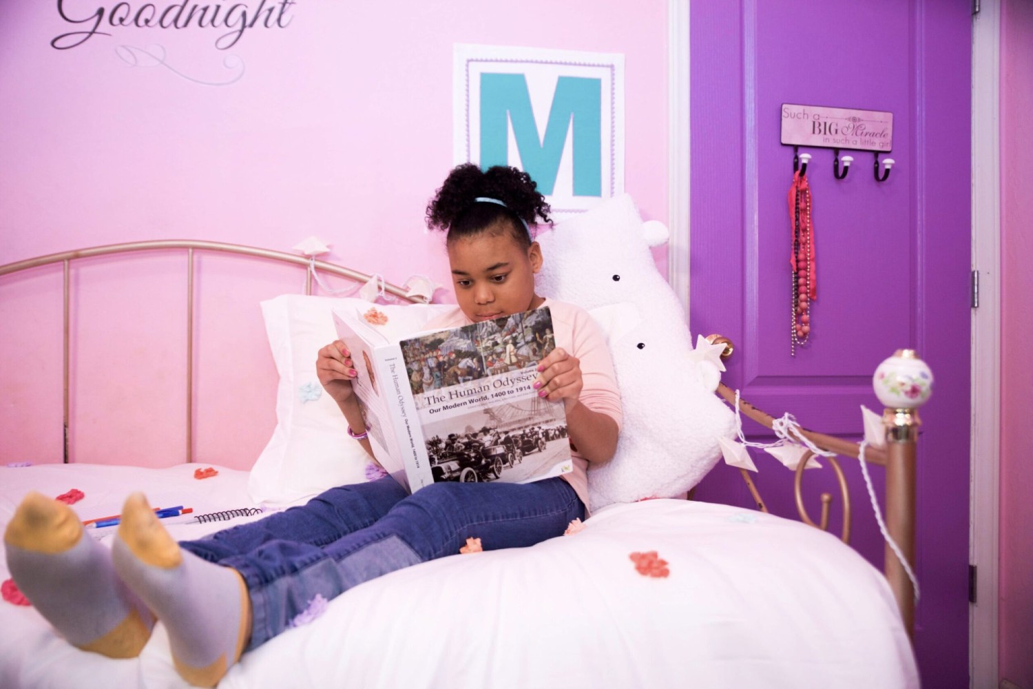 A young girl reads a history book on her bed in a pink-themed room. She looks focused and relaxed. Purple door with hanging jewelry in the background.