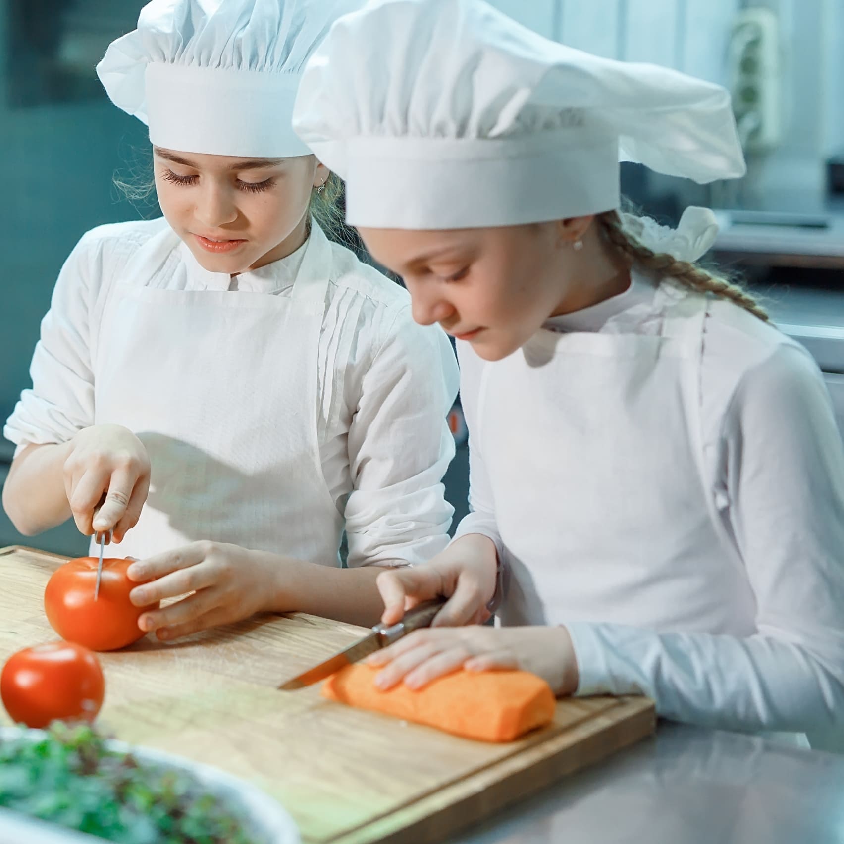 Two young chefs in white uniforms and hats are focused on chopping vegetables. One slices a tomato while the other cuts a carrot on a wooden board.
