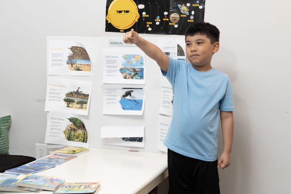 A boy in a blue shirt presents with confidence, pointing towards an animal habitats poster board. Books are spread on a table, and a solar system poster is on the wall.