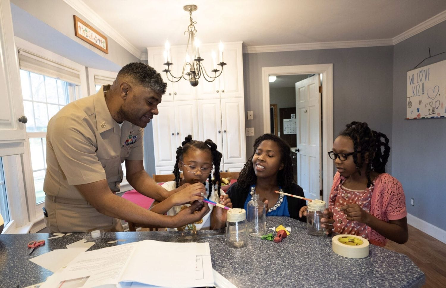 A family at a kitchen counter, engaged in a science experiment with jars and straws. The atmosphere is warm and educational, fostering learning.