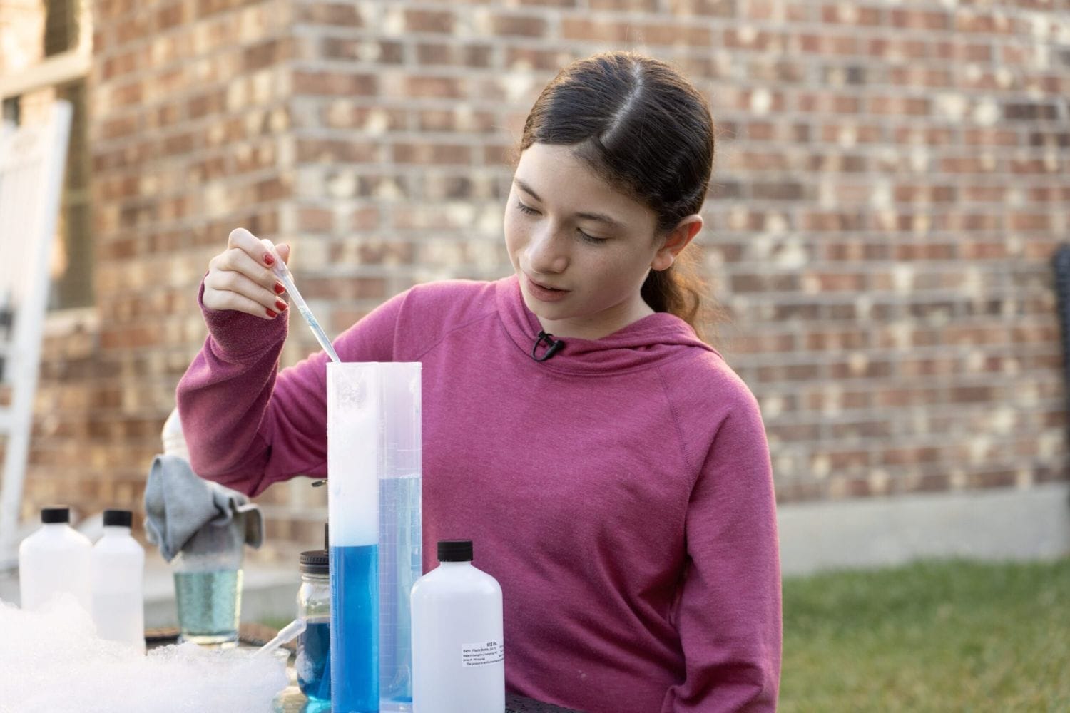 A young girl in a pink hoodie uses a dropper to add liquid to a tall cylinder filled with blue liquid. She is focused, with a brick wall behind her.