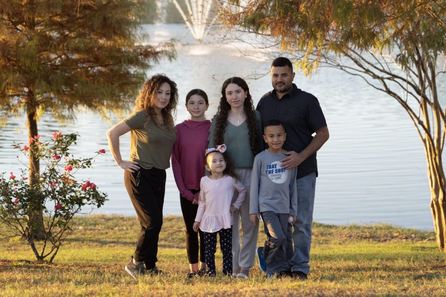 Family of six posing by a lake with blooming pink flowers and autumn trees. The setting is serene and joyful, capturing a warm, golden hour scene.