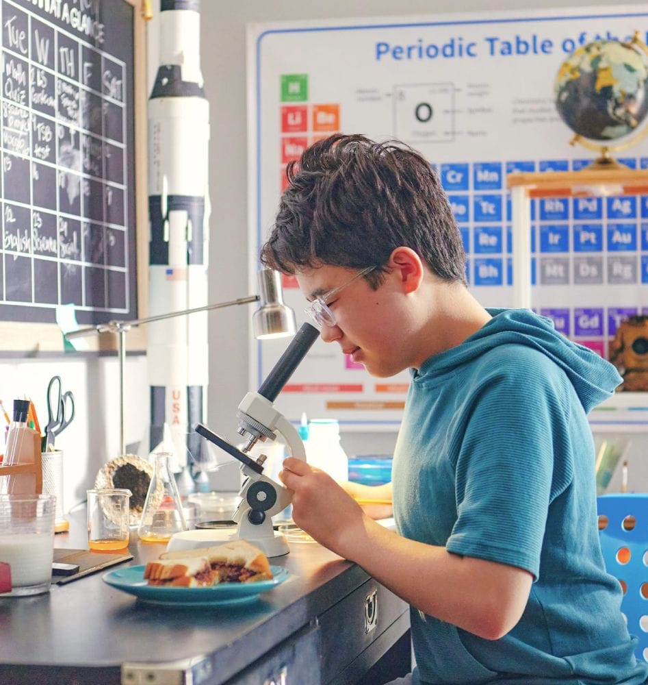 Young boy examining food under a microscope in a science-themed room, featuring a periodic table, blackboard, and model rocket, conveying curiosity.