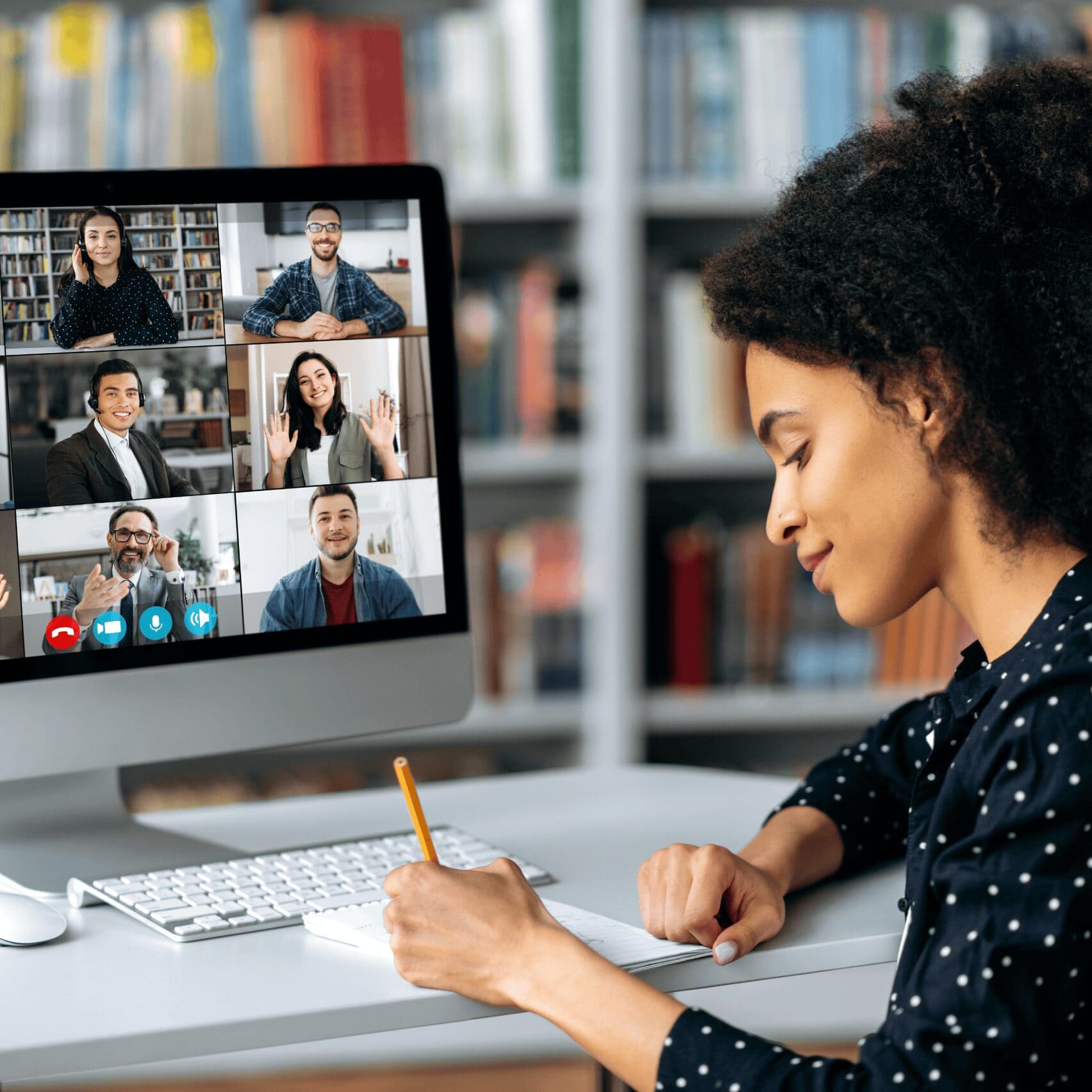 A woman takes notes while attending a video conference with six diverse participants on a computer screen, conveying focus and collaboration in a home office.