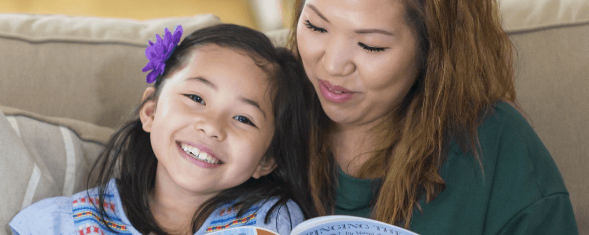 A woman and young girl sit on a couch reading a colorful book about animals. The girl smiles brightly, exuding joy and warmth.