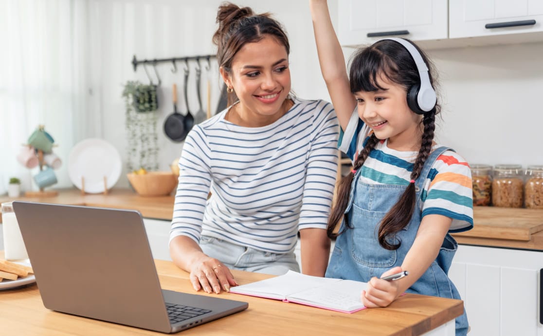A woman and a young girl are smiling in a kitchen, seated at a table with a laptop. The girl, wearing headphones, raises her hand enthusiastically.