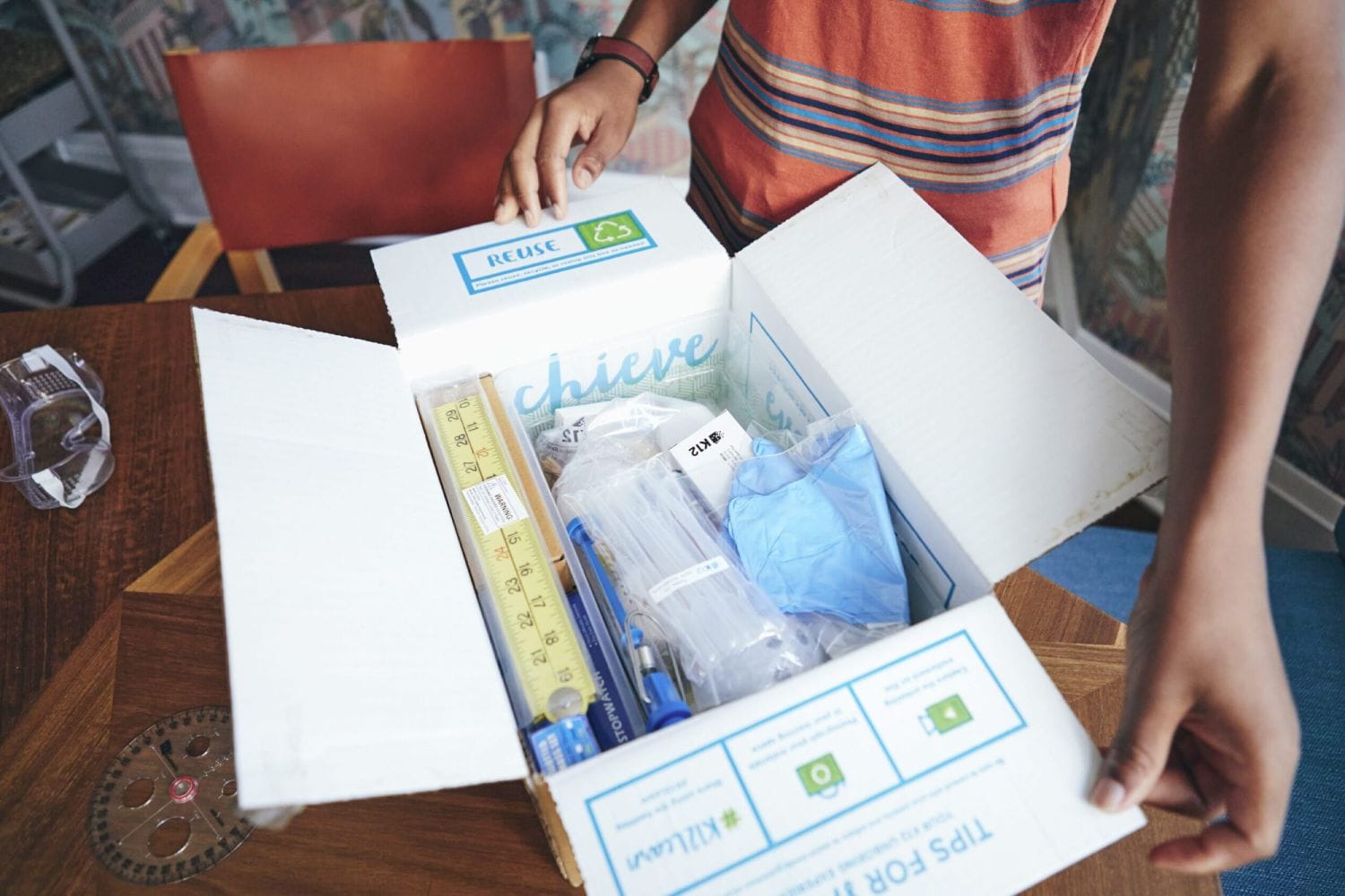 A person opens a box labeled "REUSE" containing educational supplies like rulers, gloves, and pens, set on a wooden table with a protractor.