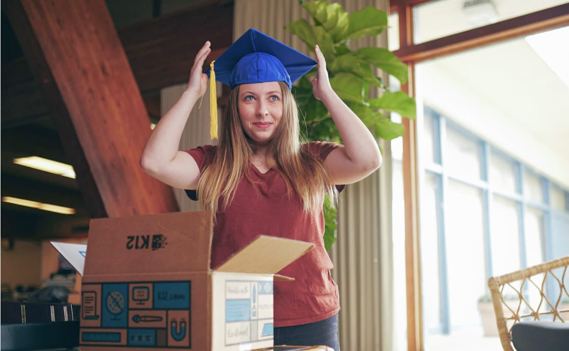 A woman with long hair, wearing a maroon shirt, adjusts a blue graduation cap. She stands indoors beside a large plant and an open cardboard box, looking pleased.