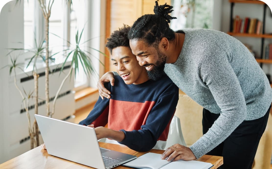 Father and son smiling at a laptop, in a cozy room with sunlight. The father leans over his son's shoulder, conveying warmth and support.