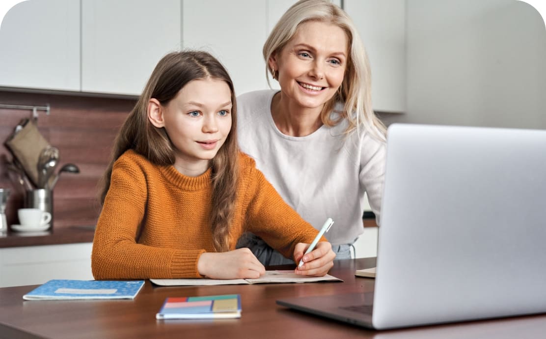 A mother and daughter smiling while looking at a laptop screen in a kitchen. The daughter writes with a pen, and notebooks are on the table. Warm, supportive atmosphere.