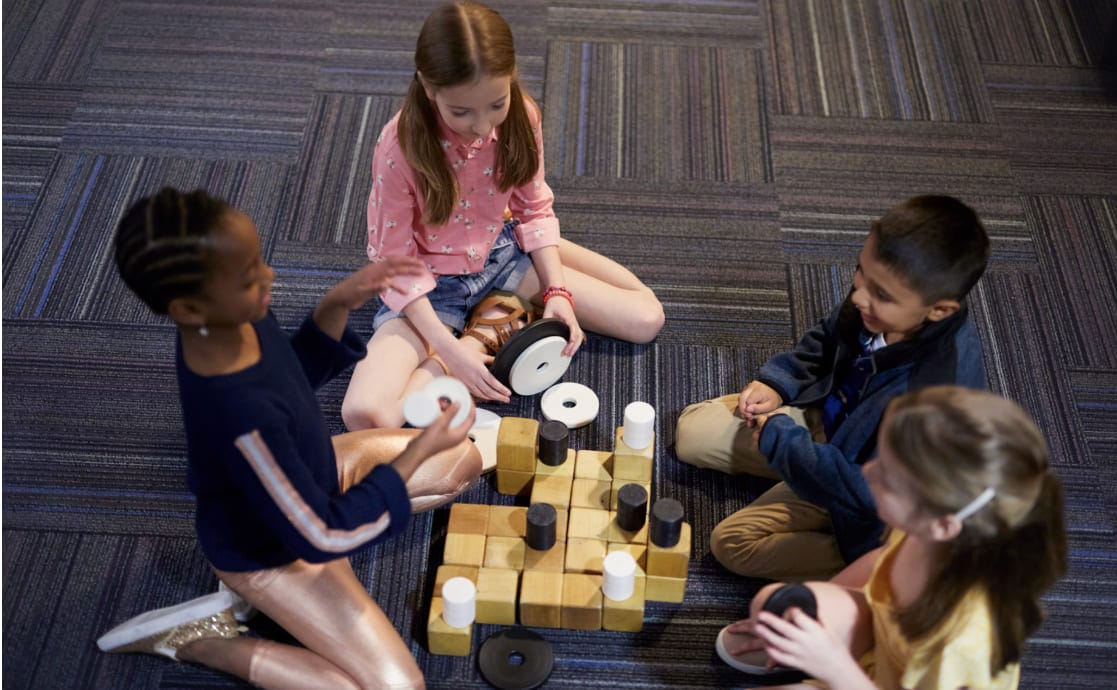 Four children sit on a carpeted floor, engaged in a board game with large disks and wooden blocks. They are focused and appear to be enjoying the game.