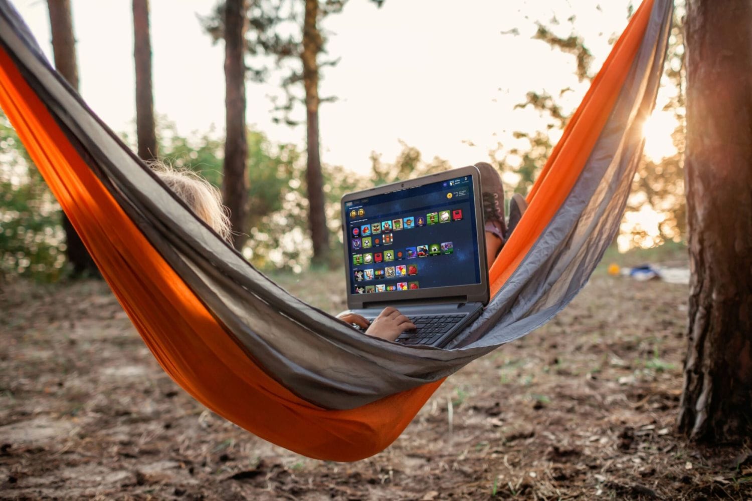 A person lounging in an orange hammock outdoors, using a laptop displaying games. Sunlight filters through trees, creating a peaceful, relaxed vibe.