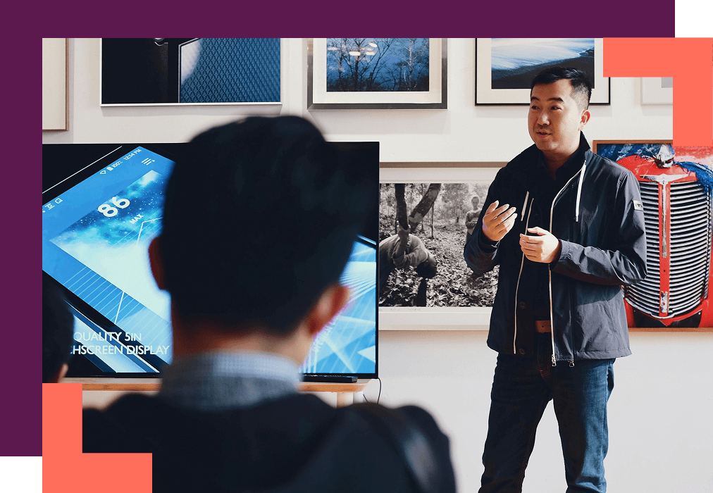 A man in a jacket presents inside a gallery-like room, with a digital screen, to an audience of one. The mood is professional.