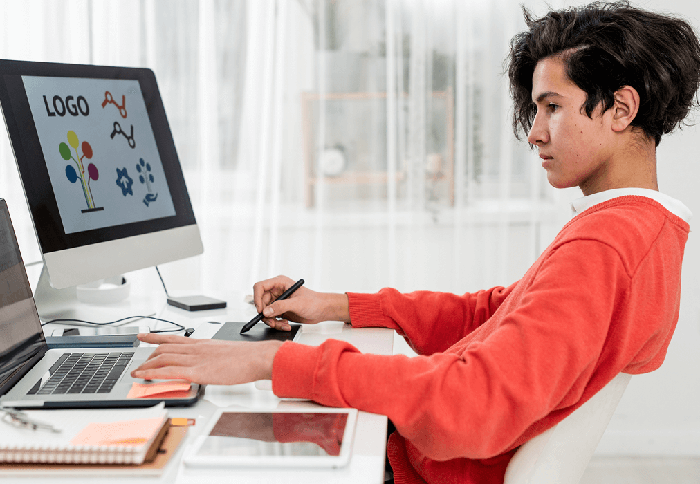 A focused person in an orange sweater works on logo designs at a desk, using a laptop and graphics tablet. The monitor displays colorful logos.