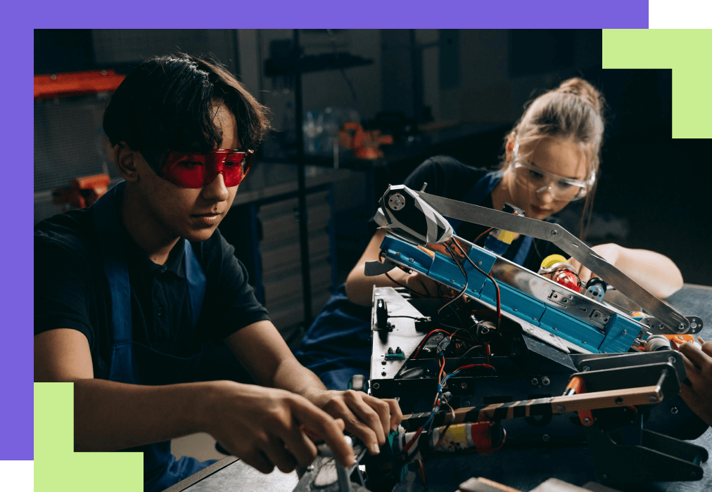 Two focused students in safety glasses work on a detailed robotic project, surrounded by wires and tools, in a workshop setting.