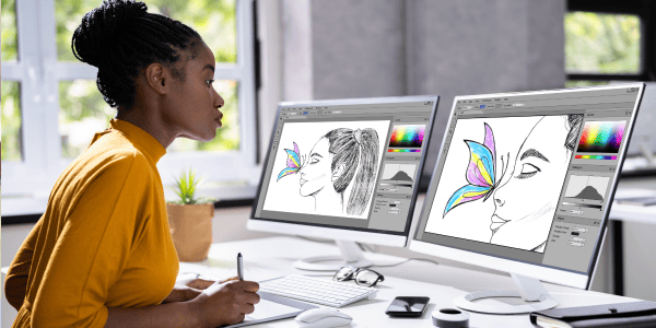 A woman in a yellow sweater works on digital illustrations of faces and colorful butterflies on dual monitors. The scene is bright and focused.