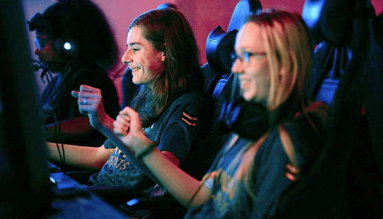 Three gamers in a dimly lit room, wearing headphones, are immersed in a game. They're smiling, raising fists in excitement, conveying a cheerful mood.