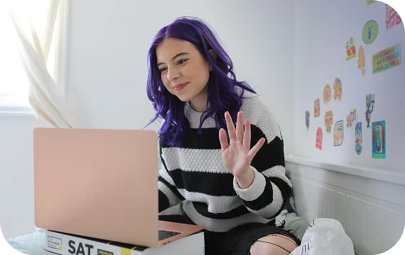 Young woman with purple hair waves at a laptop during a video call. She wears a striped sweater and sits on a bed, with colorful stickers on the wall behind.