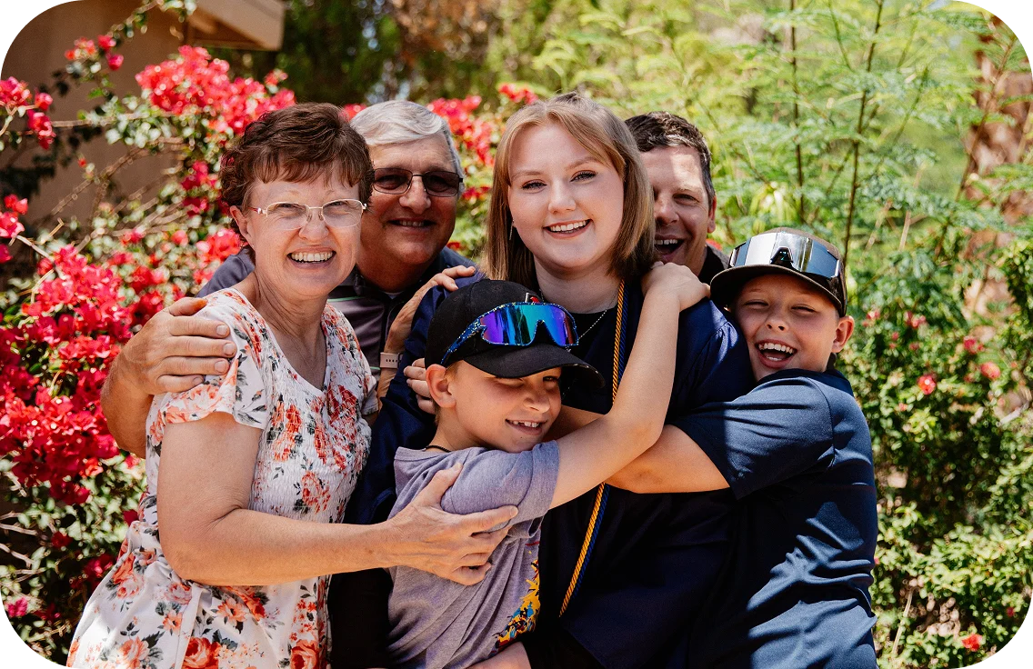 A joyful family group celebrates outdoors, with a backdrop of vibrant red flowers. They embrace and smile warmly, conveying happiness and togetherness.
