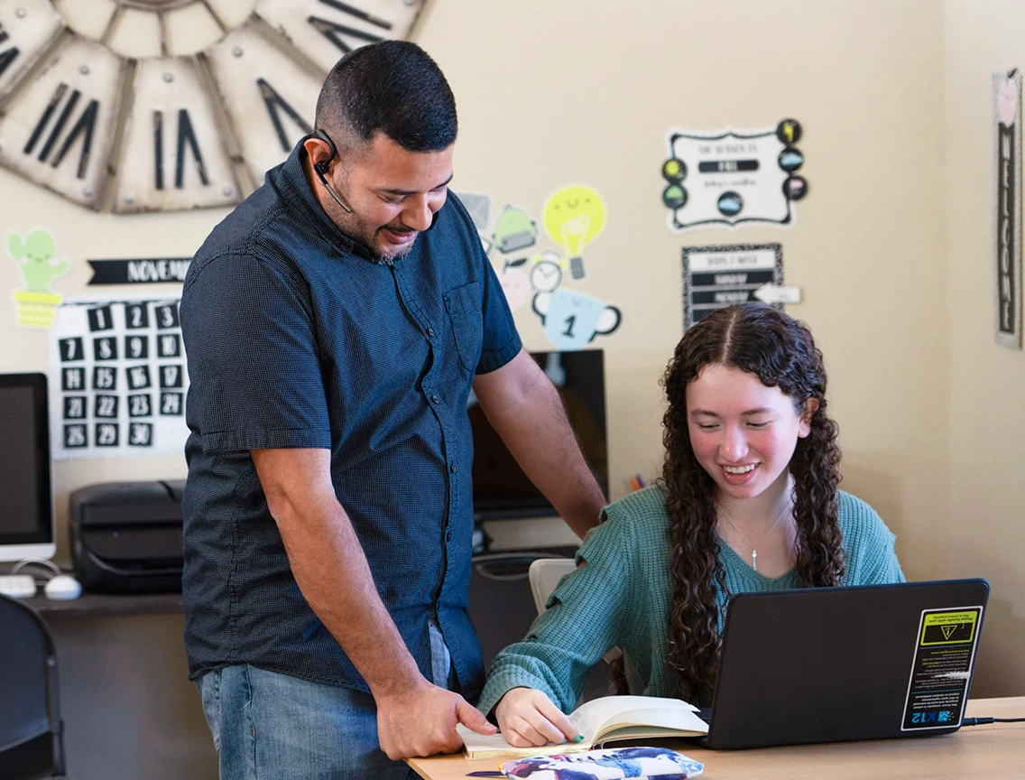 A teacher and a student are in a classroom. The teacher is smiling and leaning over a desk to help the student, who is also smiling, as she works on a laptop.